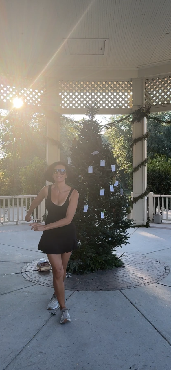 Woman in black dress and sunglasses standing on a concrete patio in front of a decorated Christmas tree during sunset.
