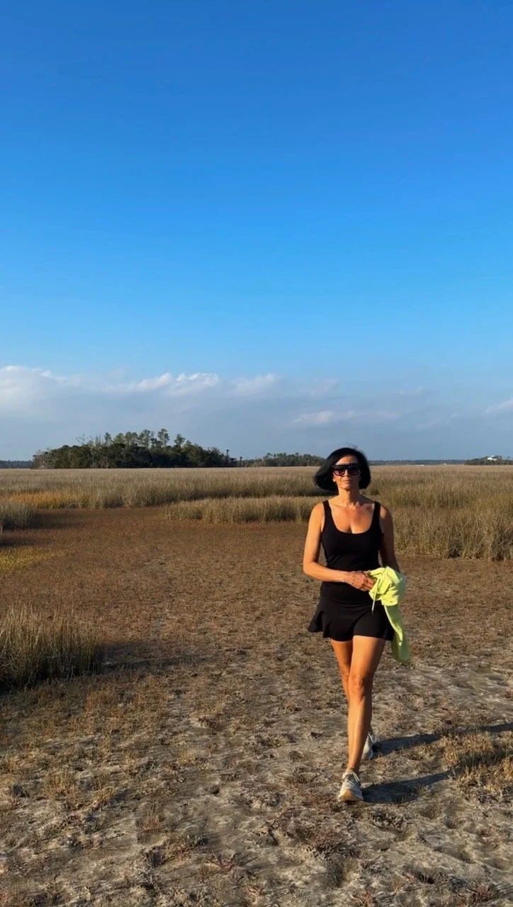 A woman with dark hair wearing sunglasses, a black tank top, and a black skirt walking on a dirt path in a grassy field under a clear blue sky, holding a yellow jacket.