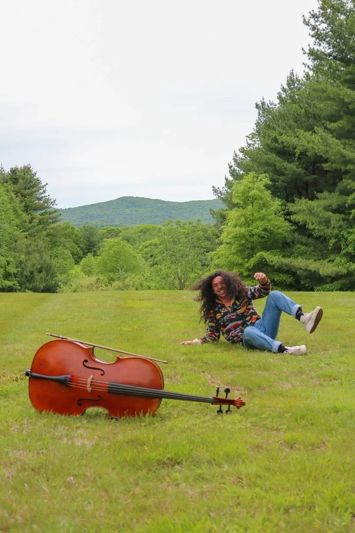 Person sitting in a grassy meadow beside a cello, with pine trees and wooded hills in the background.