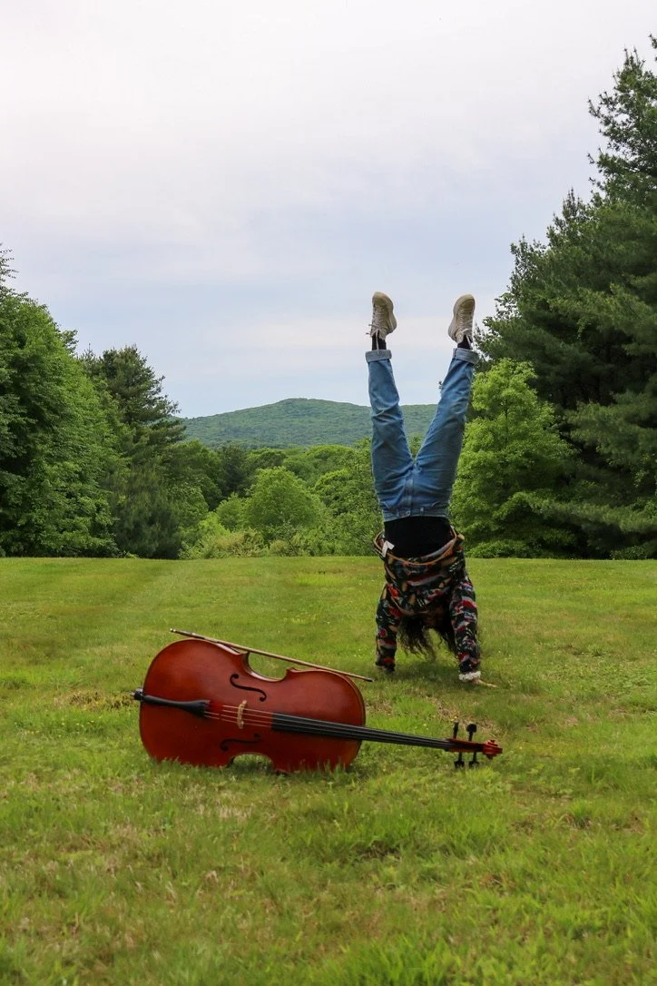 Person doing a handstand beside a cello in a grassy field with forested hills in the background