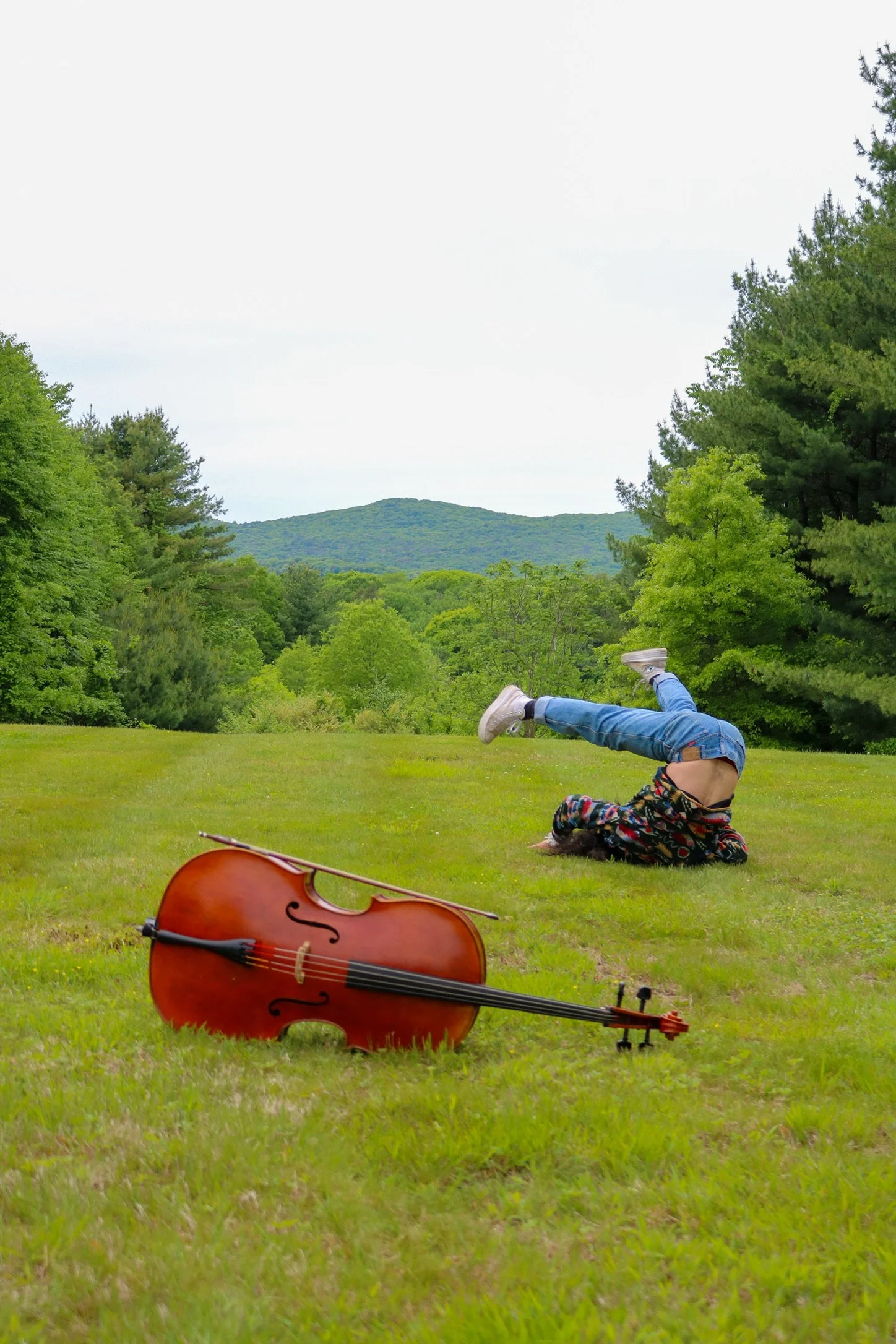 Cello lying on a grassy meadow with a person performing a floor acrobatic pose and forested hills in the background