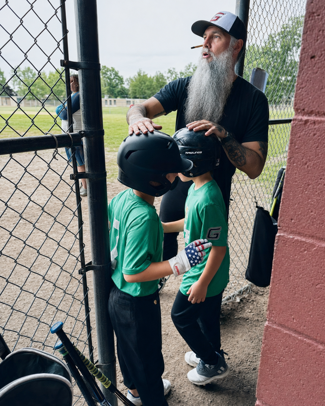 A bearded man with a baseball cap talking to two young baseball players in green jerseys, who are wearing black helmets and gloves, standing near a chain-link fence at a baseball field.