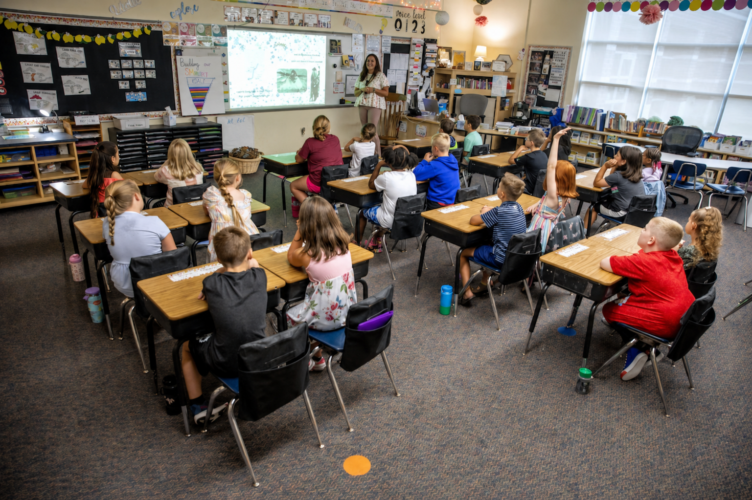 A classroom with students sitting at desks, facing a teacher at the front. Students have notebooks and water bottles. The classroom has large windows, a whiteboard, and educational posters on the walls.