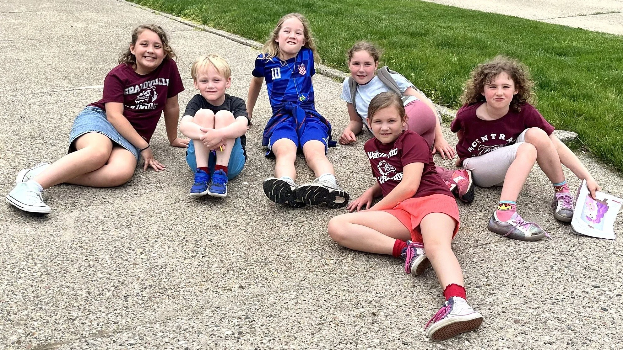 A group of seven children relaxing on a sidewalk. They are smiling and sitting or lying on the ground, with green grass beside them.