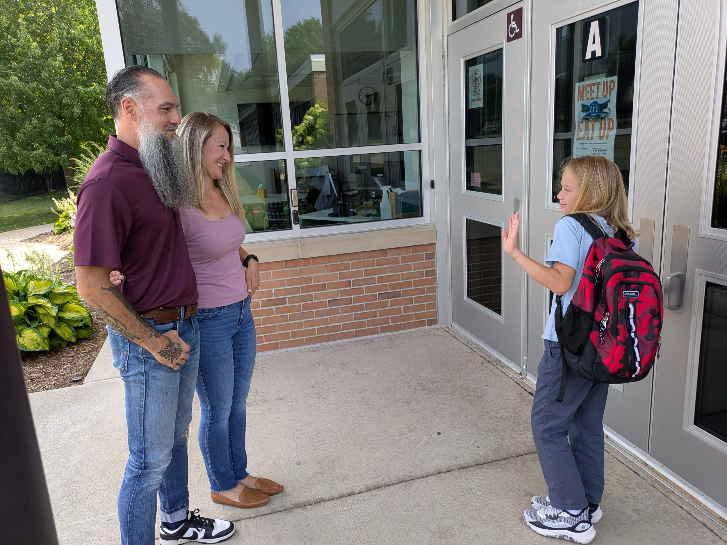 A girl in a blue shirt and backpack greets a woman and man outside a building with large glass windows, near a gray door with an 'A' sign, a disability parking sign, and various stickers, including a 'Meet Up and Eat Up' poster.