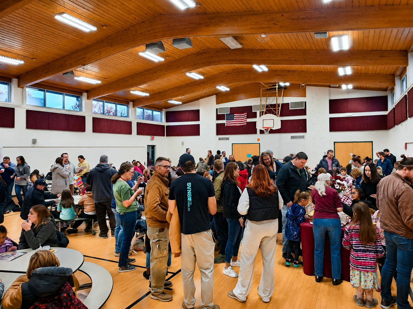 Crowd of people inside a gymnasium, some sitting at tables and some lining up around tables, with a basketball hoop, American flag, and red soundproof panels on the walls.