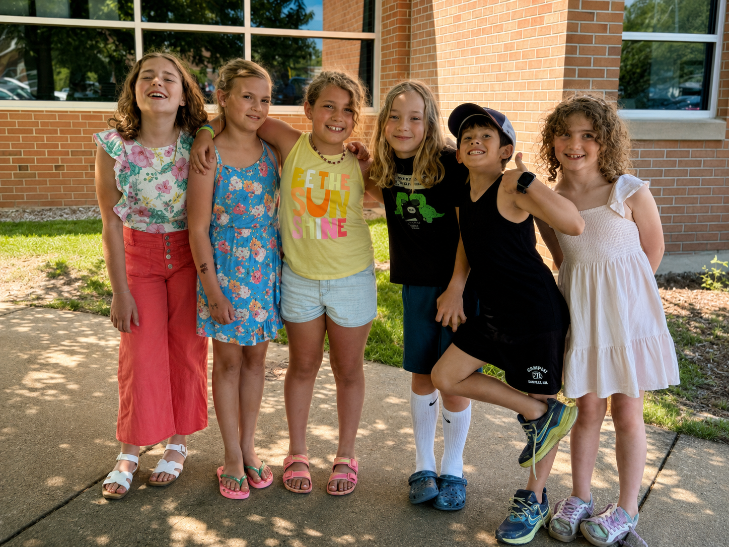 Six children standing outside, smiling and posing together, in front of a brick building with large windows, on a sunny day.