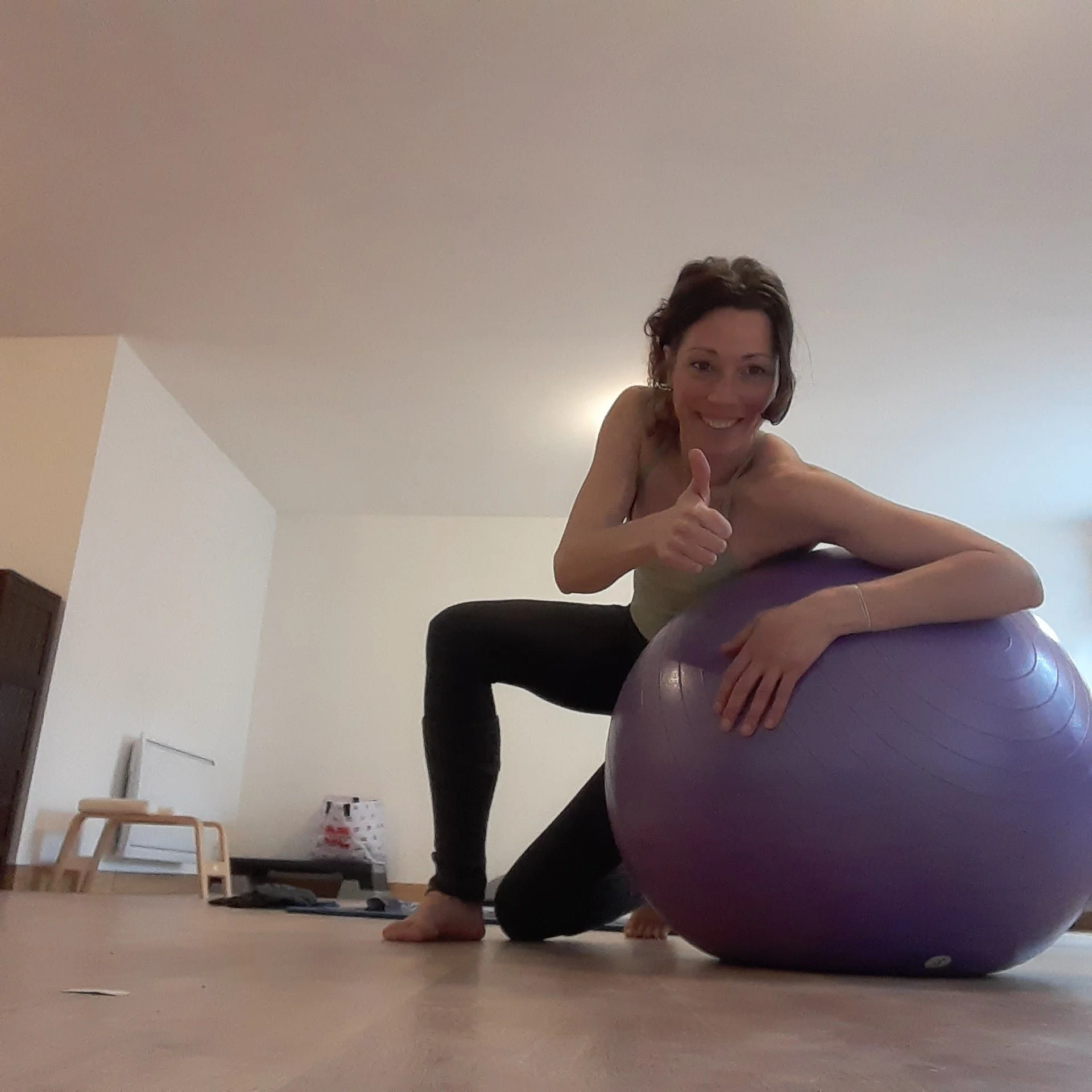 Une femme souriante faisant un exercice de yoga ou de fitness sur un tapis, avec un ballon d'exercice violet, dans une pièce intérieure avec un sol en bois clair et quelques meubles en arrière-plan.