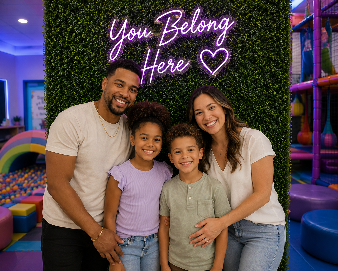 A family of four smiling together at an indoor kids' play area with a green wall and neon sign that says 'You Belong Here' with a heart. The background features colorful play equipment and a ball pit.