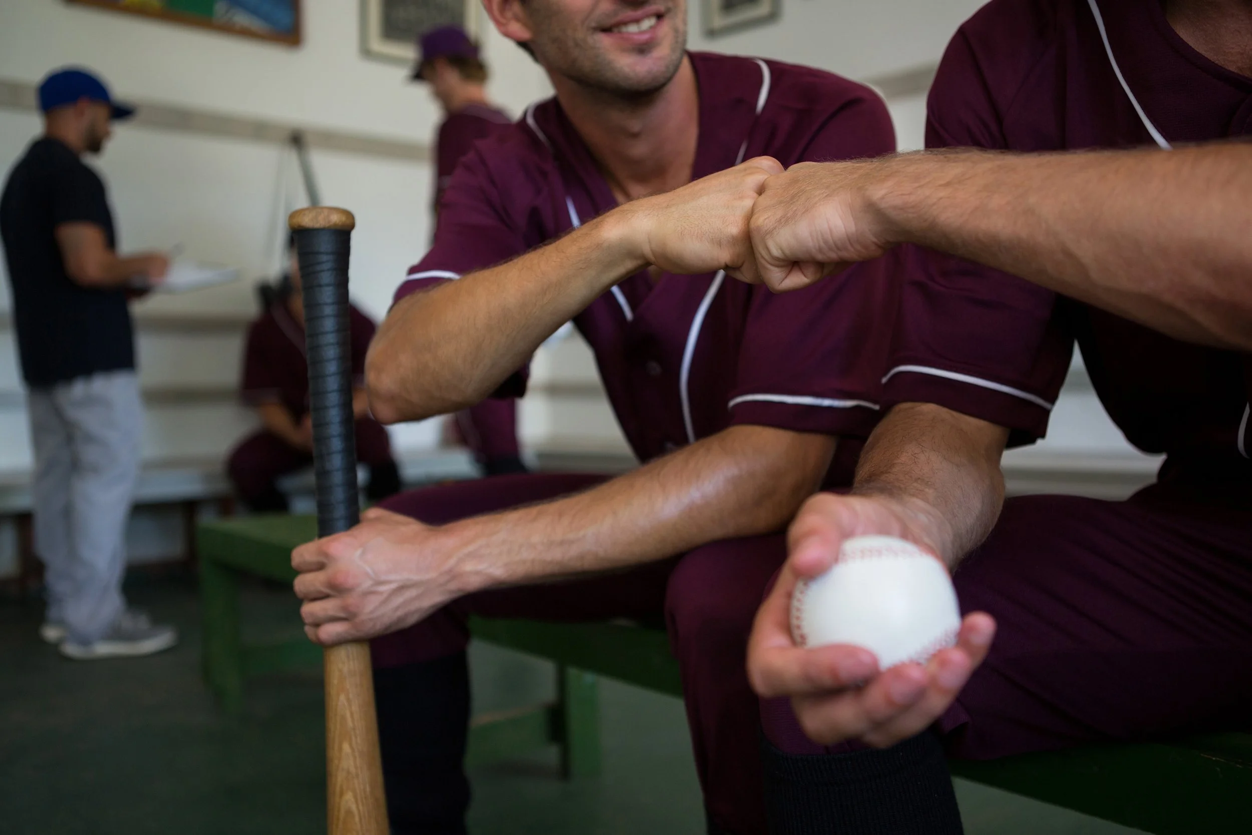 Two men in matching maroon shirts engaging in a handshake, seated at a table with baseball gloves and a ball, in a room with other people and framed pictures on the wall.
