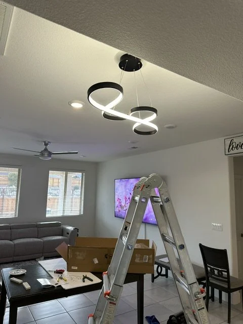 Modern living room with a unique black and white swirl ceiling light fixture, a television on the wall, a gray sofa, and a chair. A ladder and a cardboard box are also visible.