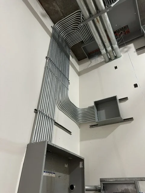 A corner of an unfinished room showing electrical conduit pipes running along the walls and ceiling, with an empty electrical panel box on the wall.