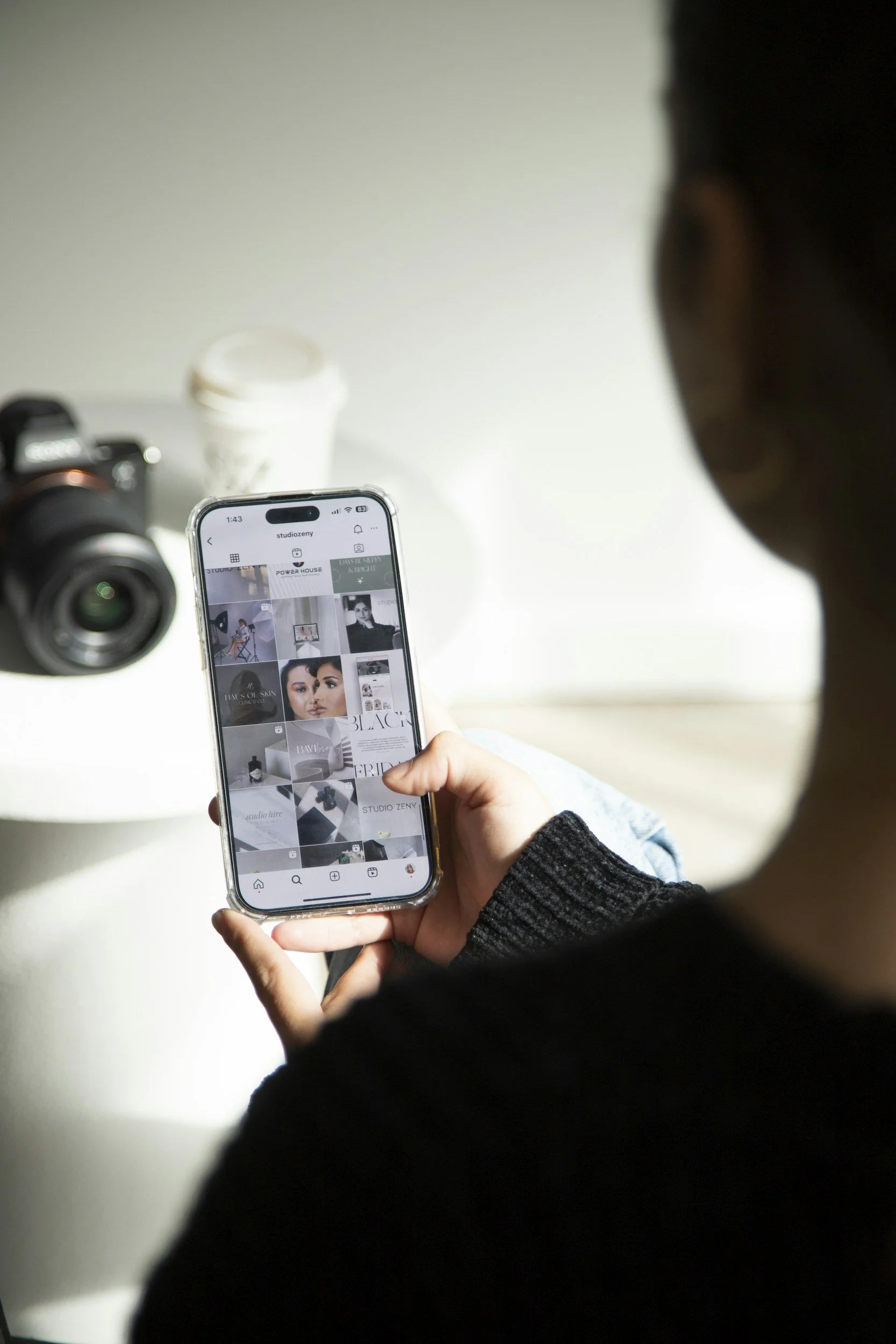Person viewing a phone screen with a grid of images, sitting near a camera and a white cup on a white surface.
