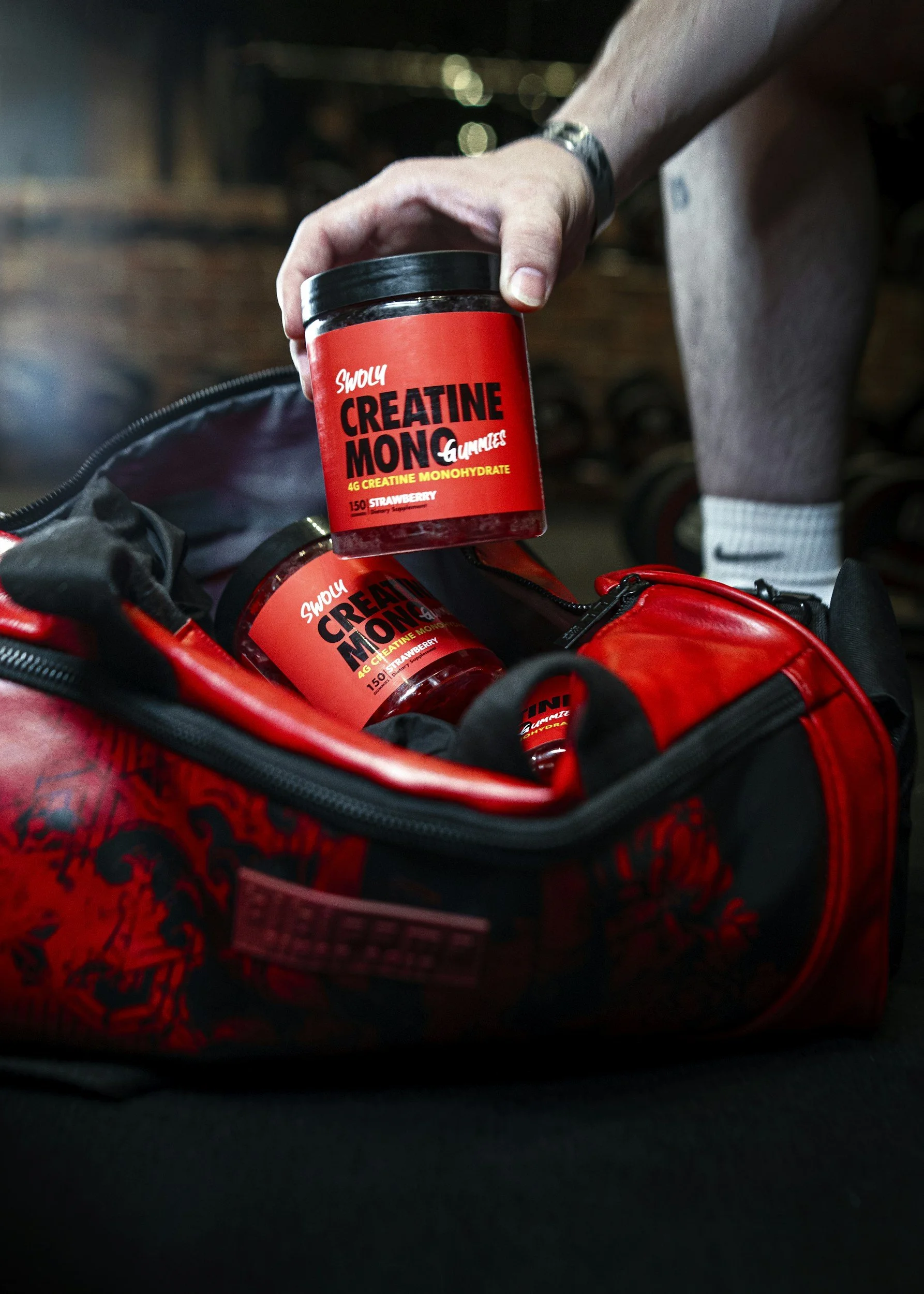 A person holding a container labeled 'Swoly Creatine Monohydrate' with strawberry flavor in a red and black bag, inside a gym bag.
