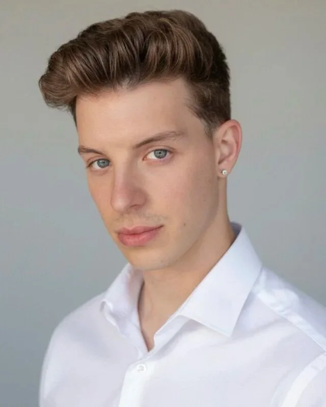 A young man with styled brown hair, blue eyes, and wearing a white collared shirt and a small earring in his left ear, looking at the camera against a plain gray background.