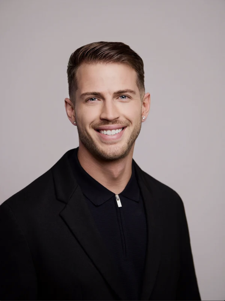 Portrait of a smiling young man with short brown hair, light stubble, earrings, wearing a black blazer and black zip-up polo shirt against a plain light gray background.