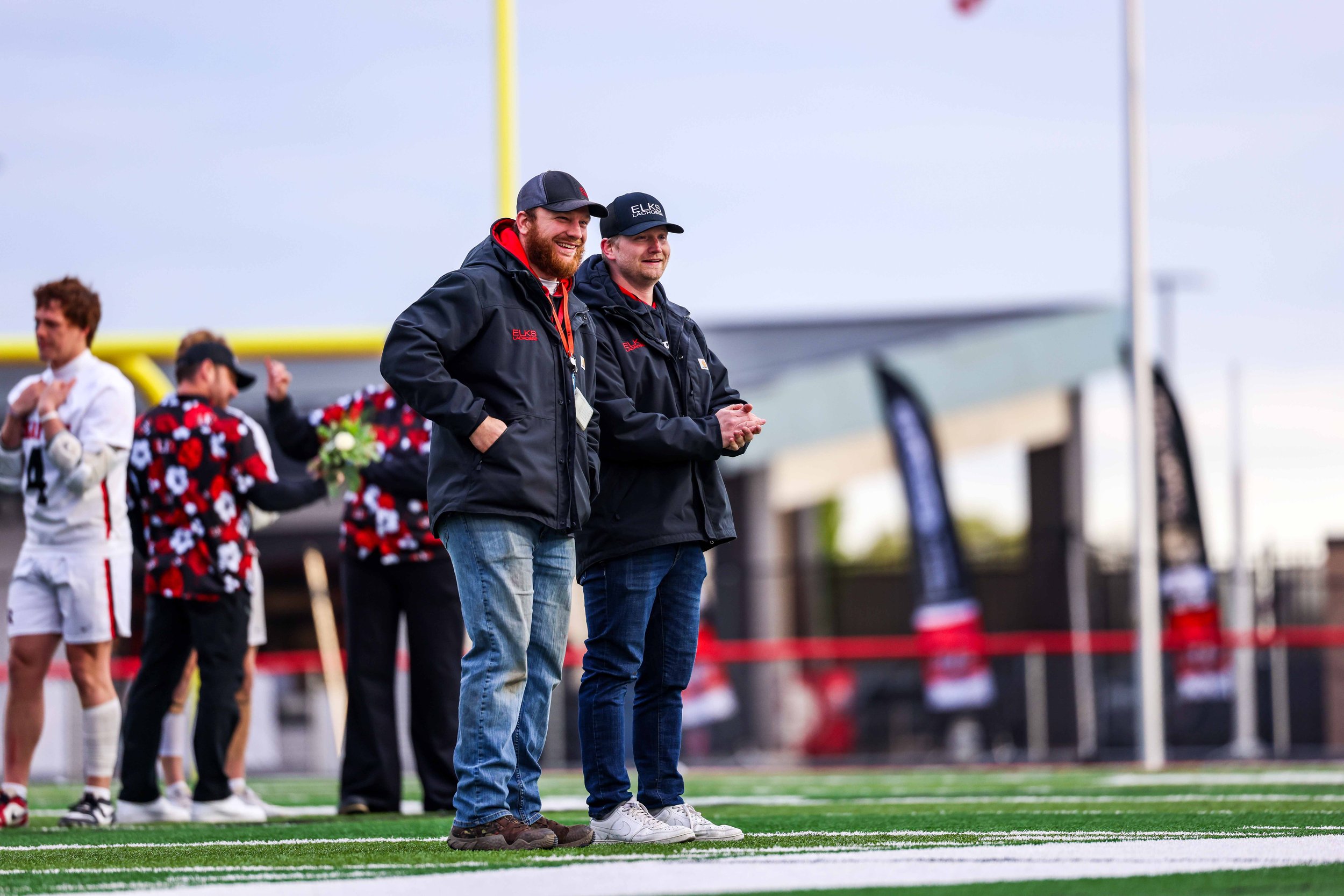 Two men standing on a football field during a game, with other players in the background. The man on the left has a beard, wearing jeans and a dark jacket, smiling. The man on the right is wearing jeans and a cap, looking forward. Both are standing n