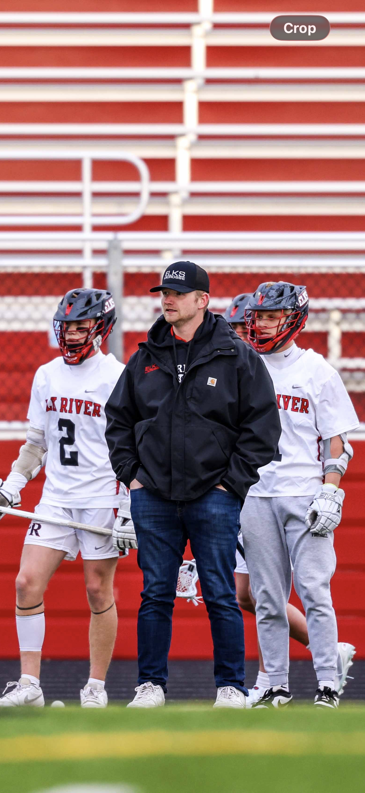 Lacrosse coach and two players standing on a field. The players are wearing helmets and lacrosse gear, with jerseys reading 'LA RIVER' and numbers 2 and 1. The coach is wearing a black jacket and cap. They are standing on the sidelines with a red sta