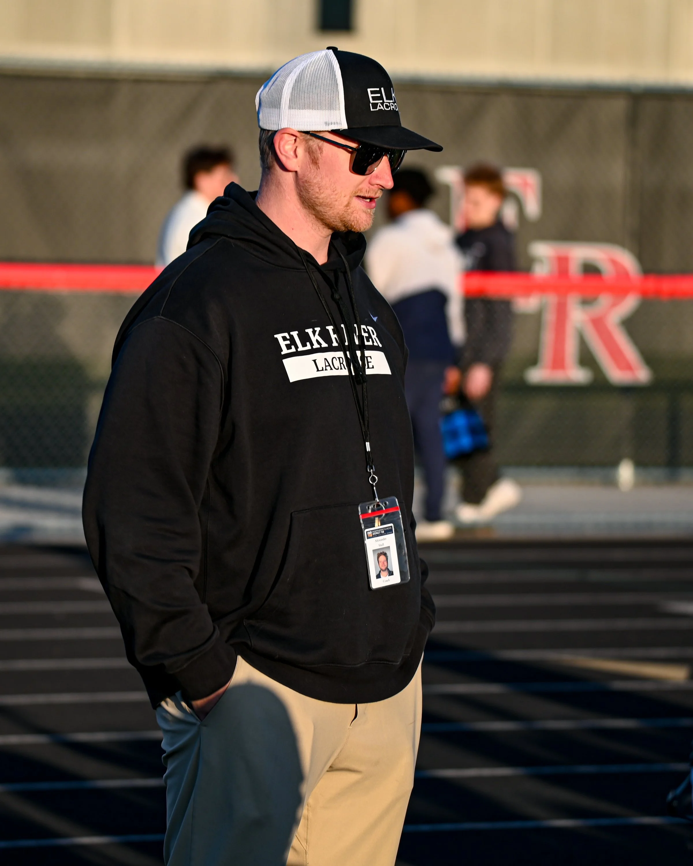 A young man wearing a black hoodie with white text, beige pants, sunglasses, and a black and white cap, standing outdoors with his hands in his pockets. He has a lanyard with a badge around his neck. There are people blurred in the background behind 
