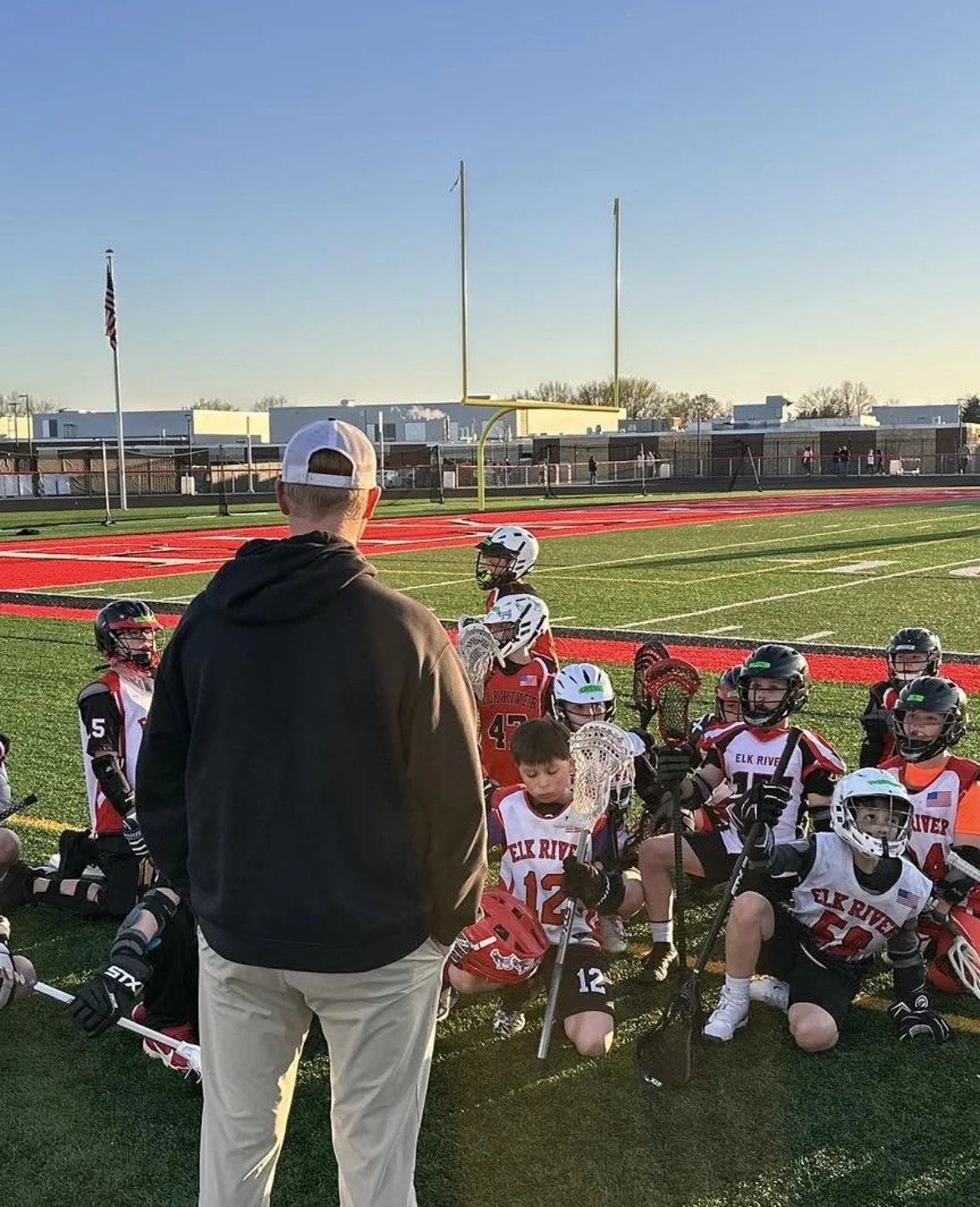 Youth lacrosse team sitting on the field listening to coach during practice or game on a football field with red track, goal post, and school buildings in background.