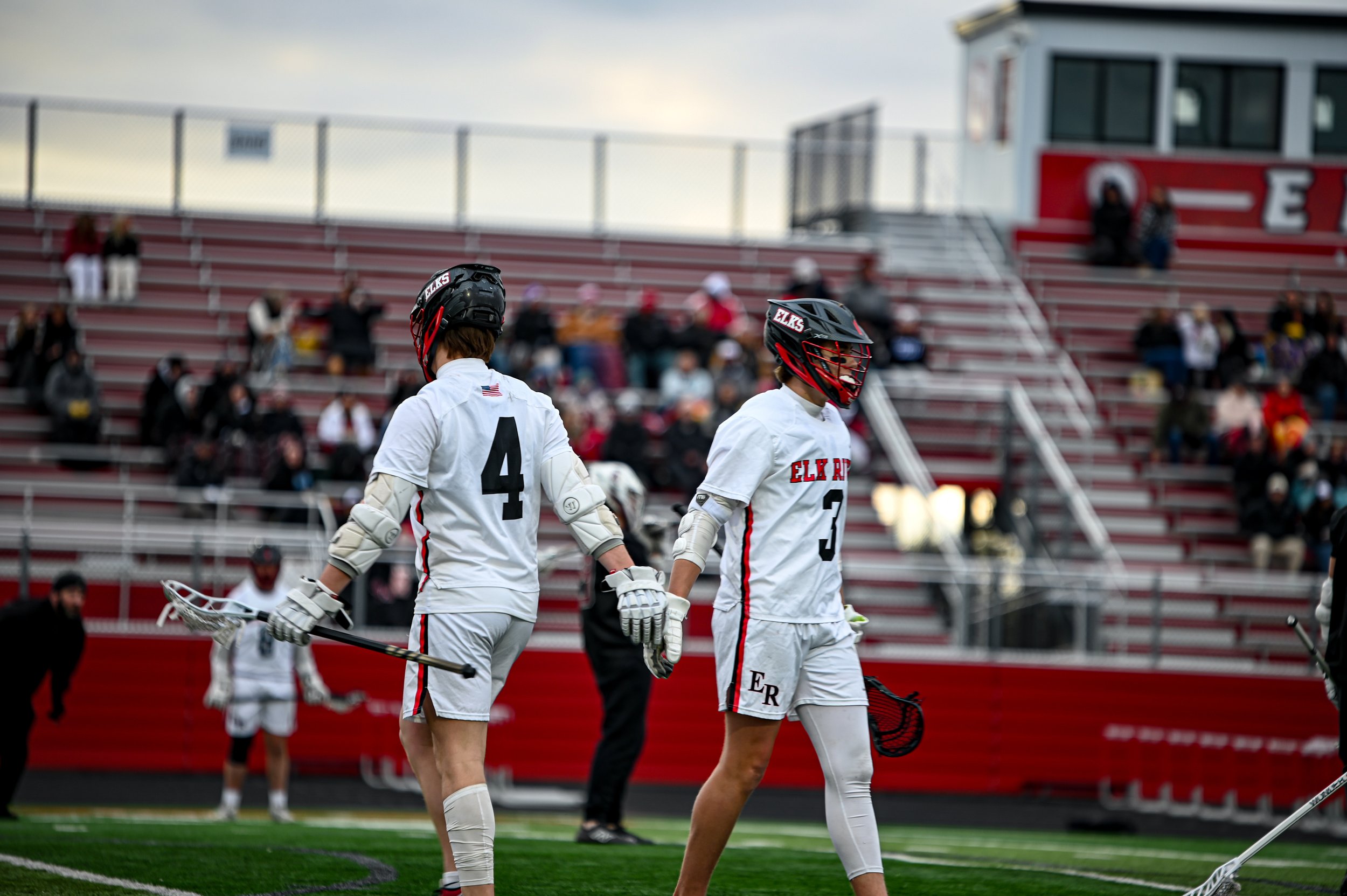 Two lacrosse players in white jerseys and black helmets walking on a sports field, with a red bleacher and spectators in the background.