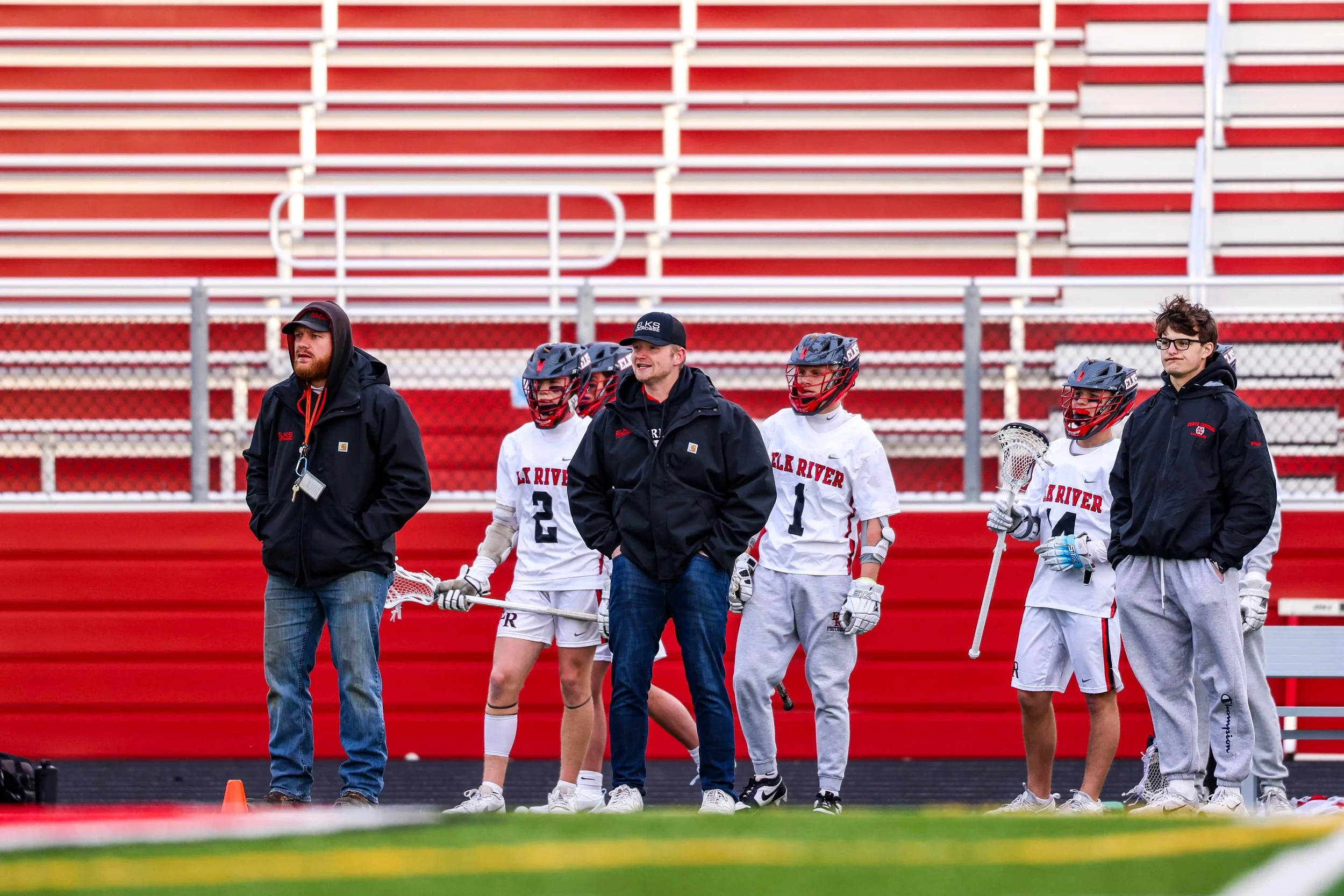 Lacrosse players and coaches standing on a sports field against red bleachers, some players wearing helmets and holding lacrosse sticks.