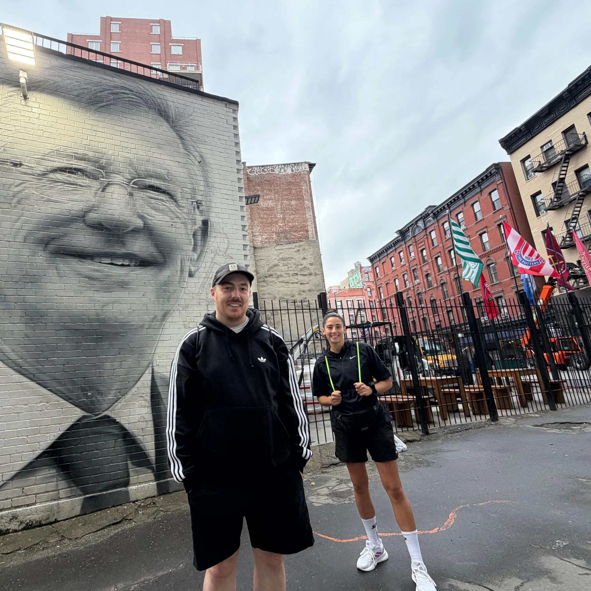 Two young people, a man and a woman, standing outdoors in an urban area near a graffiti-covered wall and colorful flags. The man is wearing a black jacket, black shorts, and a cap. The woman is dressed in black athletic wear and holding a jump rope.