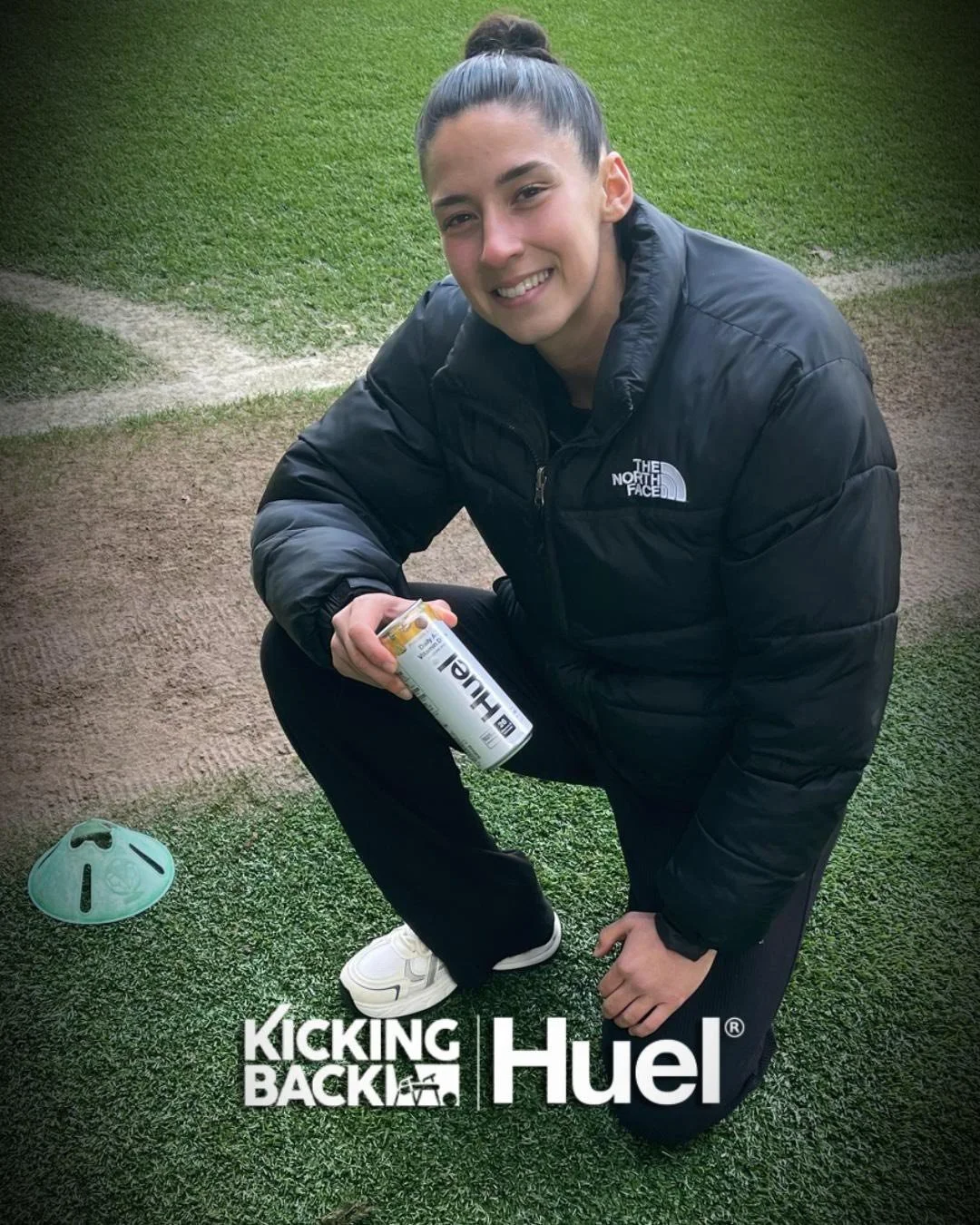 A young woman kneeling on a sports field, smiling, wearing a black North Face jacket, white sneakers, and holding a can of Huel. There is a green tuning fork-shaped object on the ground beside her with the logos "Kicking Back" and "Huel".