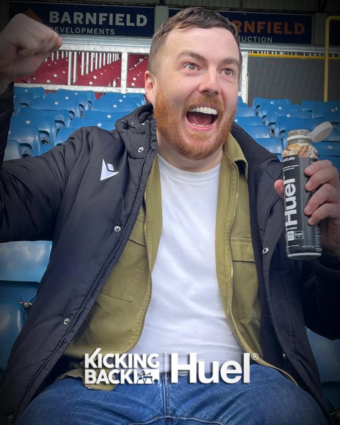A man with a reddish beard and short hair sitting in stadium seating, smiling excitedly with an open mouth, holding a can of Fuel energy drink in his right hand, raising his left fist in celebration.