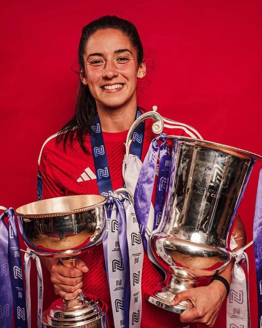 A female athlete in a red sports jersey holding two large silver trophies and wearing multiple medals around her neck, standing against a red background.