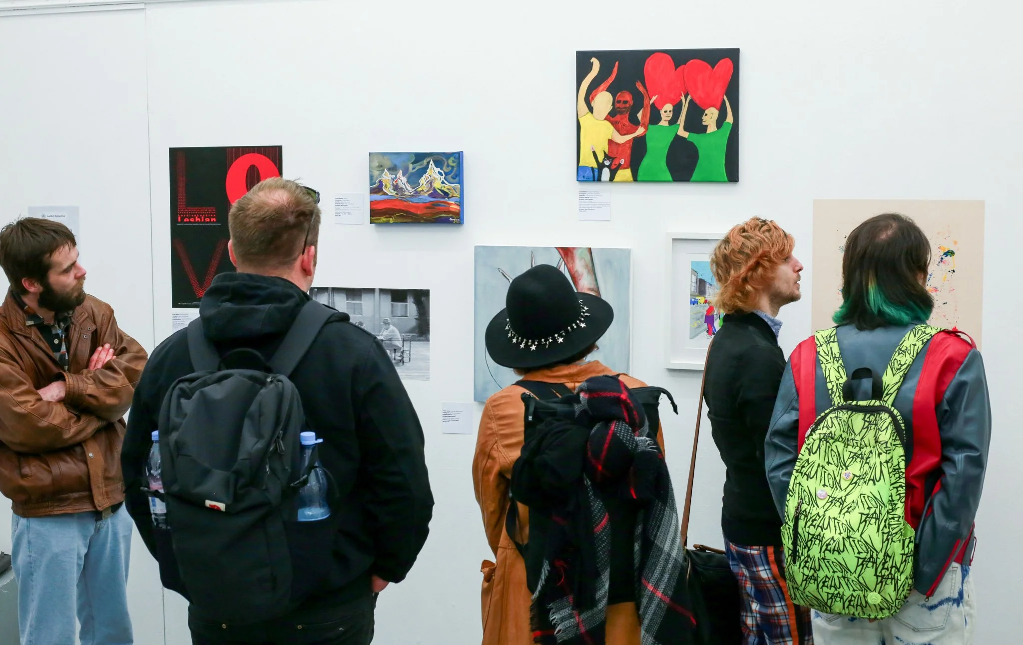 People viewing artwork in a gallery, with various paintings displayed on the white wall, including colorful abstract and figurative art.
