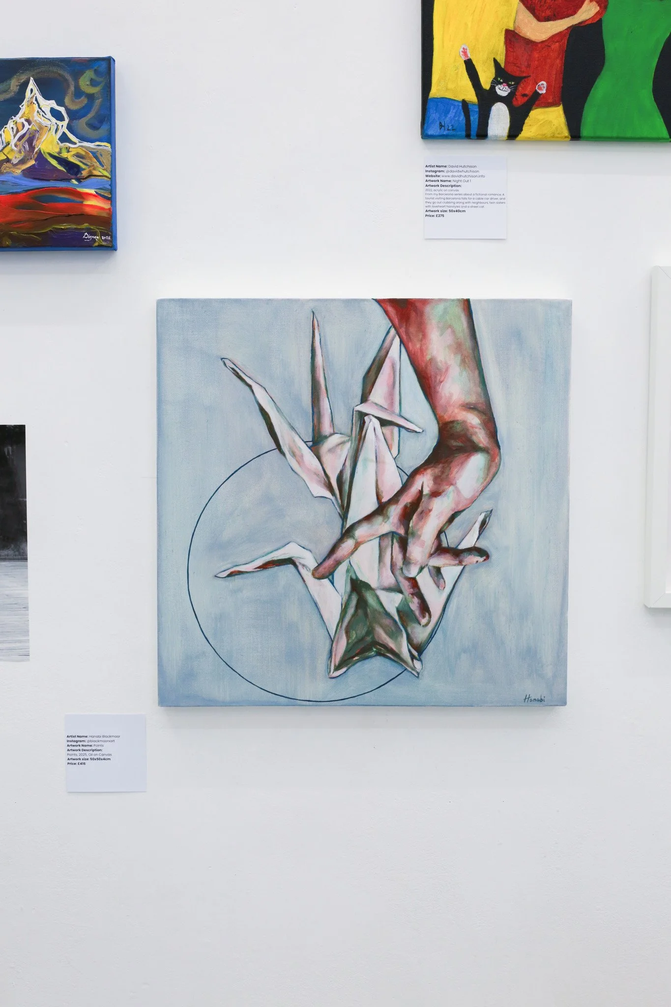 Paintings displayed on a white gallery wall, including a watercolor of a hand and paper crane, and others featuring colorful abstract designs and a black cat, each with informational placards.