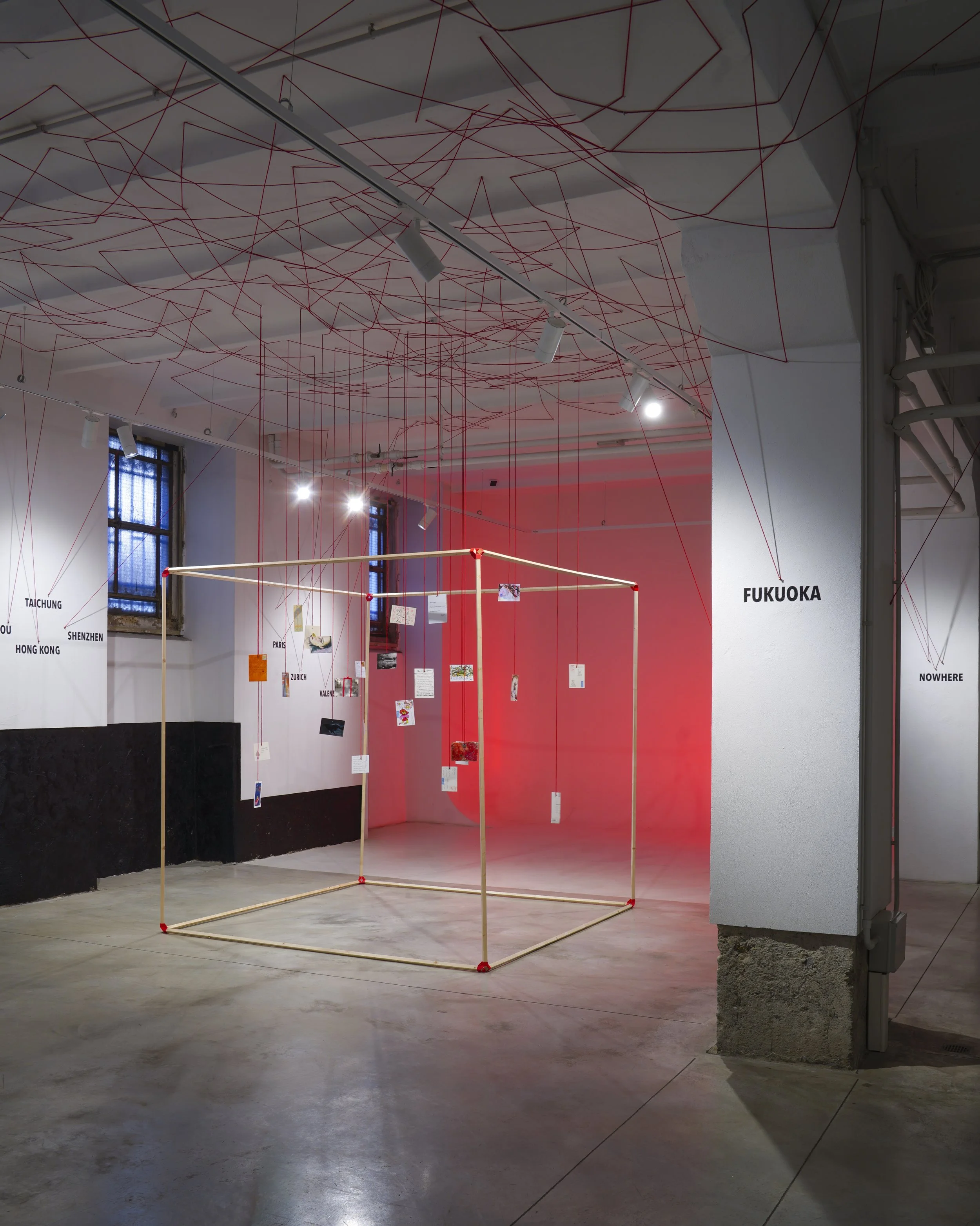 An art installation with red string creating an intricate web on the ceiling, with several hanging notes or photographs below, in a gallery space with windows and a red-lit background.
