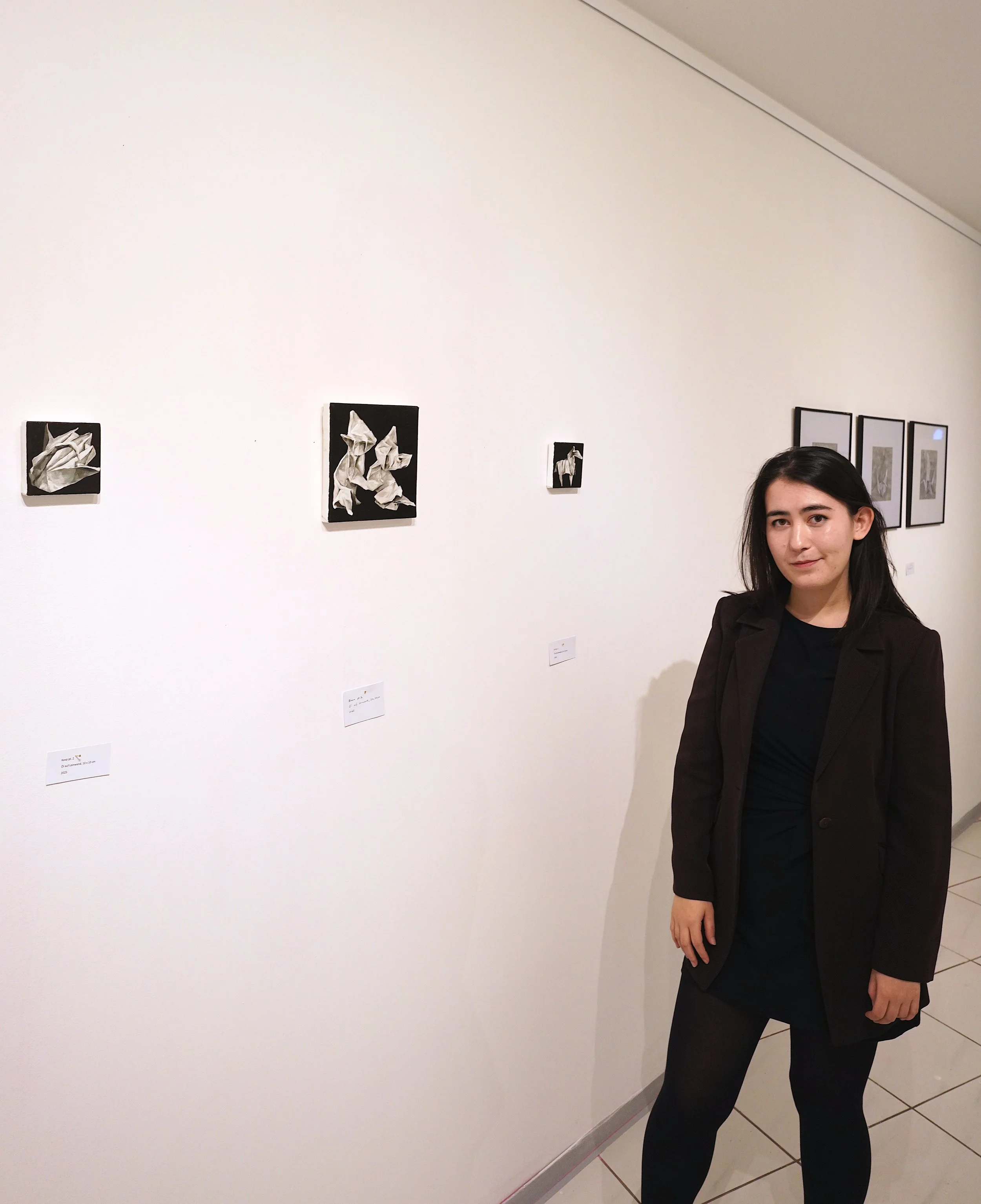 Woman with shoulder-length dark hair wearing a black blazer and black pants standing in an art gallery next to framed artworks on a white wall.