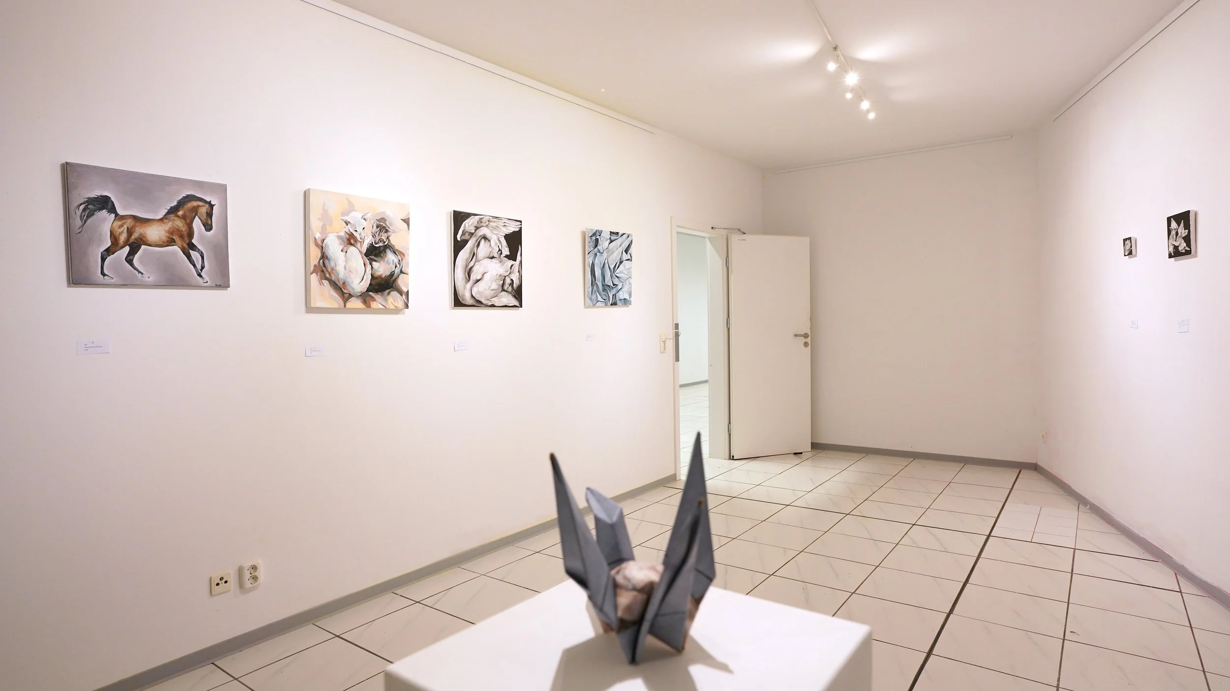 An art gallery with white walls displaying several paintings, including a horse, abstract designs, and human figures. In the foreground, a handcrafted sculpture resembling a rabbit is placed on a white pedestal.