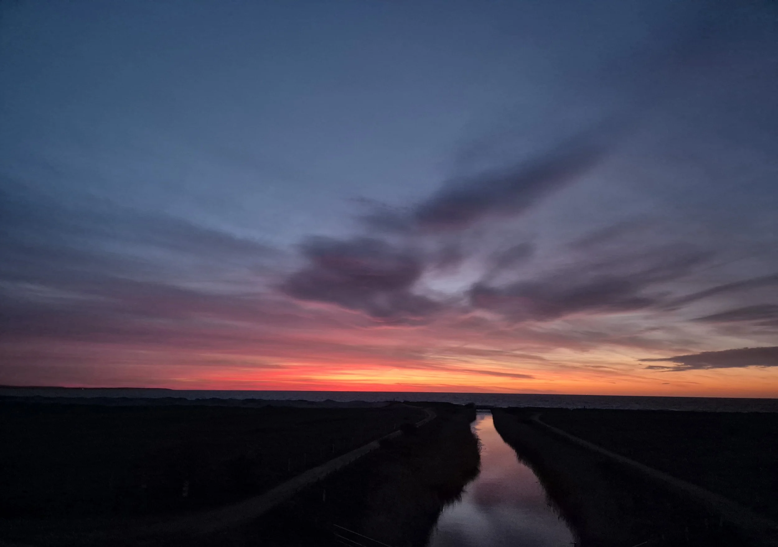 Ein farbenfroher Sonnenuntergang über einer Wasserstraße mit Spiegelung der Wolken am Himmel.