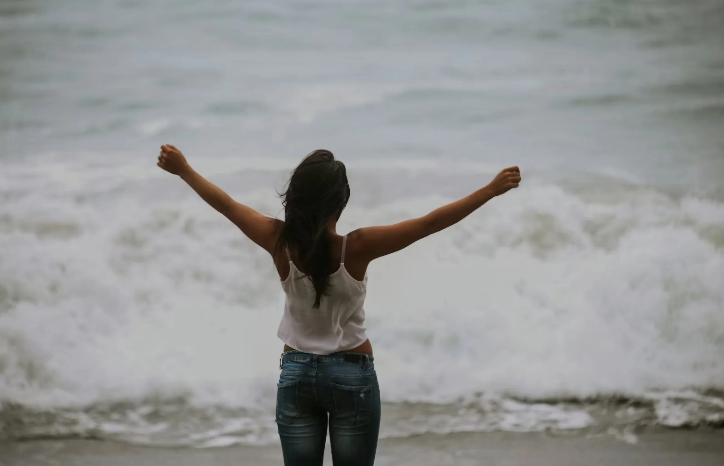 Junge Frau steht am Meer mit ausgebreiteten Armen und genießt die Meeresbrise.
