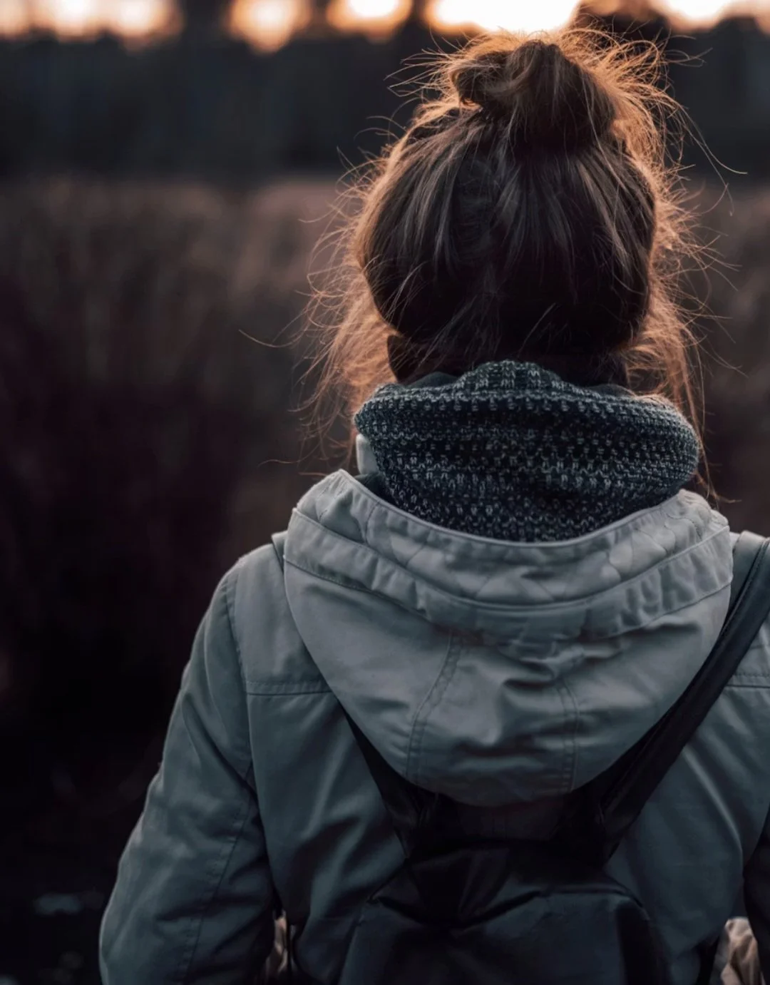 Eine Person mit braunen Haaren in einem Dutt, trägt eine hellgraue Jacke und einen dunkelgrauen Pullover, schaut auf eine dunkle Landschaft bei Sonnenuntergang.