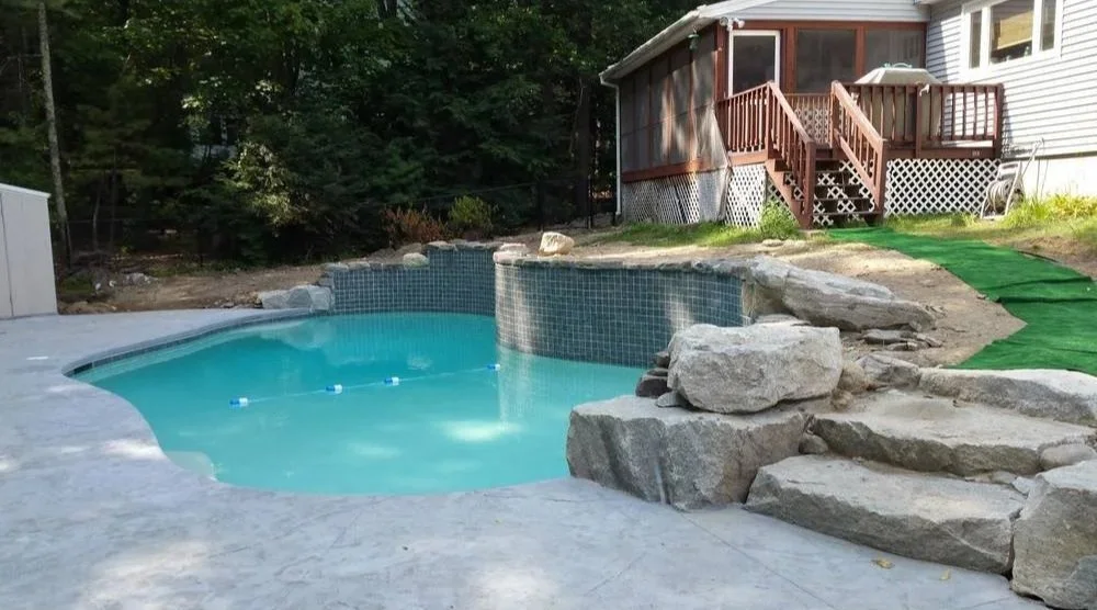 A backyard with a small pool surrounded by rocks, a house with a screened porch, and a green artificial turf path leading to the porch stairs.