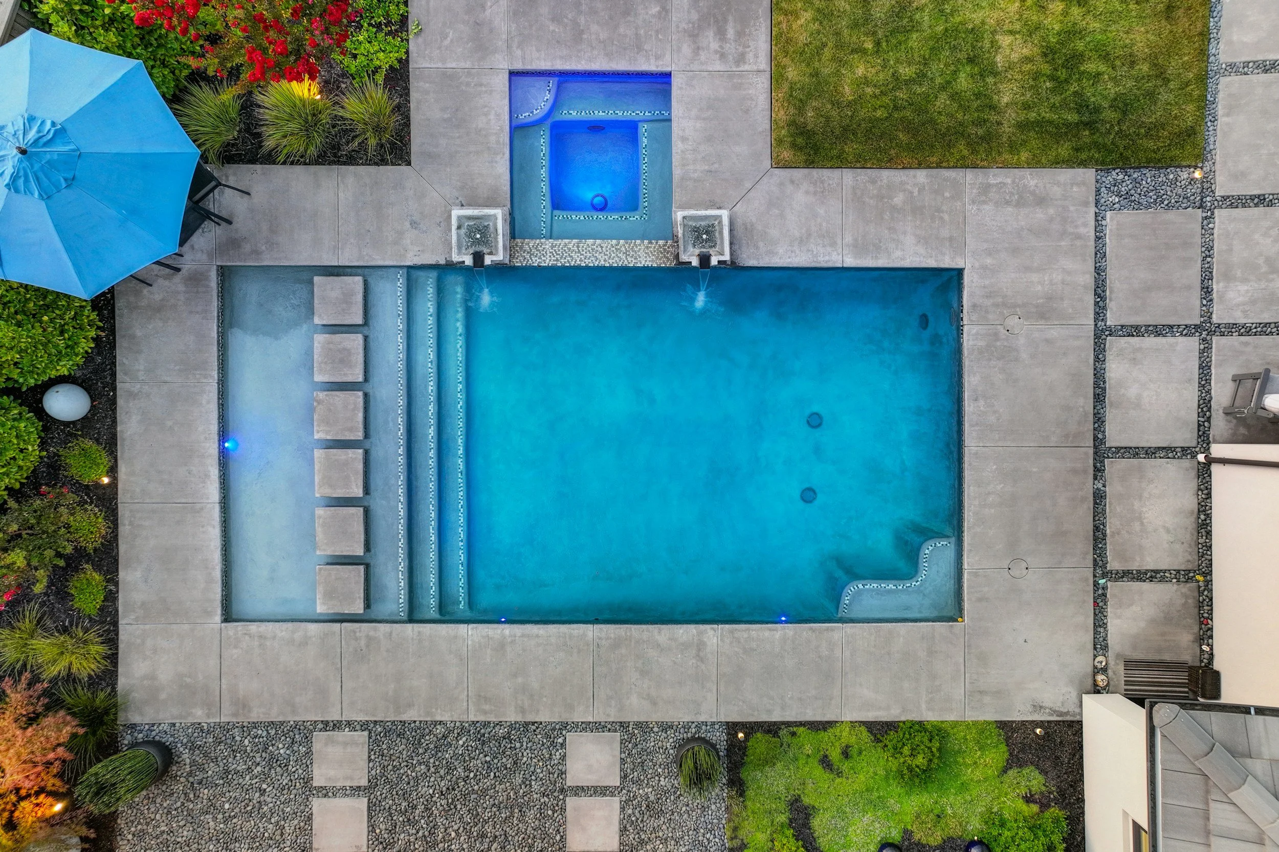 An aerial view of a backyard with a swimming pool, hot tub, patio, umbrella, chairs, grass, and landscaped garden.