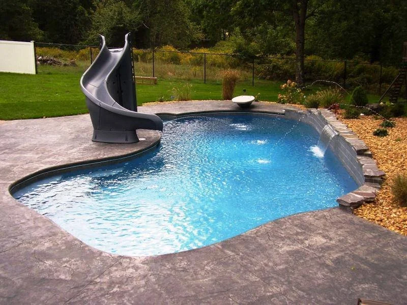 Backyard swimming pool with a water slide and a small ledge, surrounded by a concrete deck and landscaped yard with trees and plants.