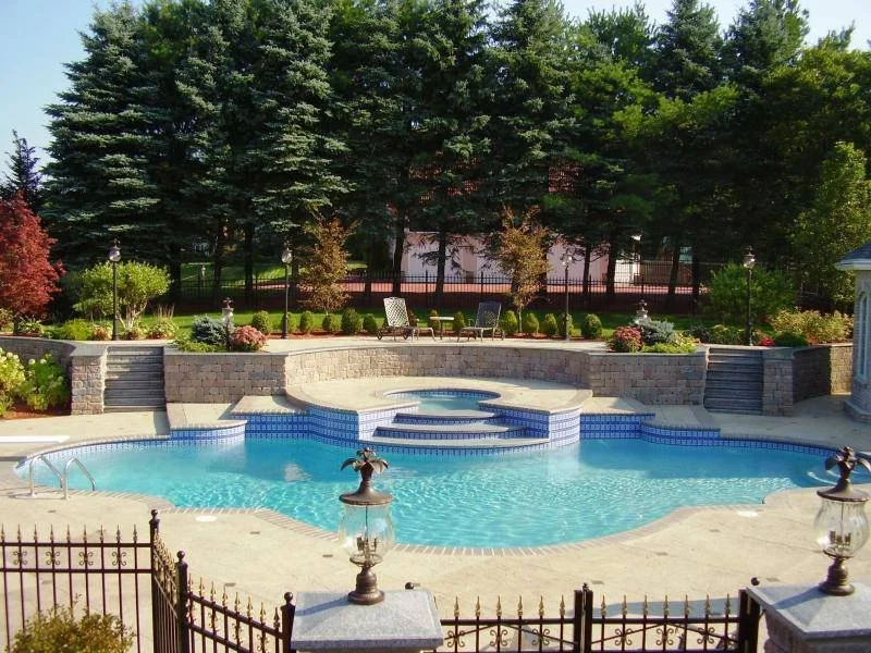 A backyard swimming pool with steps leading into it, surrounded by a decorated stone patio and garden, with trees and outdoor chairs in the background.