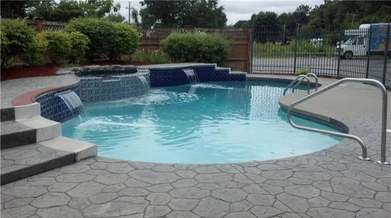 Backyard swimming pool with built-in spa and waterfall feature, enclosed by a black metal fence, with pool steps and a ladder, surrounded by a stamped concrete patio and greenery in the background.