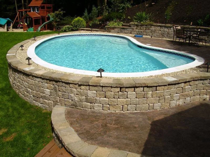 Round in-ground swimming pool with a stone border, surrounded by a paved deck and a grassy yard with trees and outdoor furniture in the background.