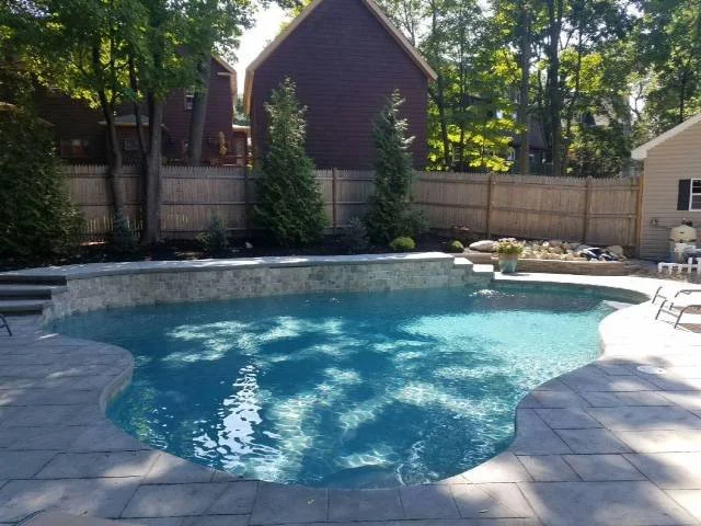 Backyard swimming pool with a stone patio, surrounded by a wooden fence and tall trees, with a purple house in the background.