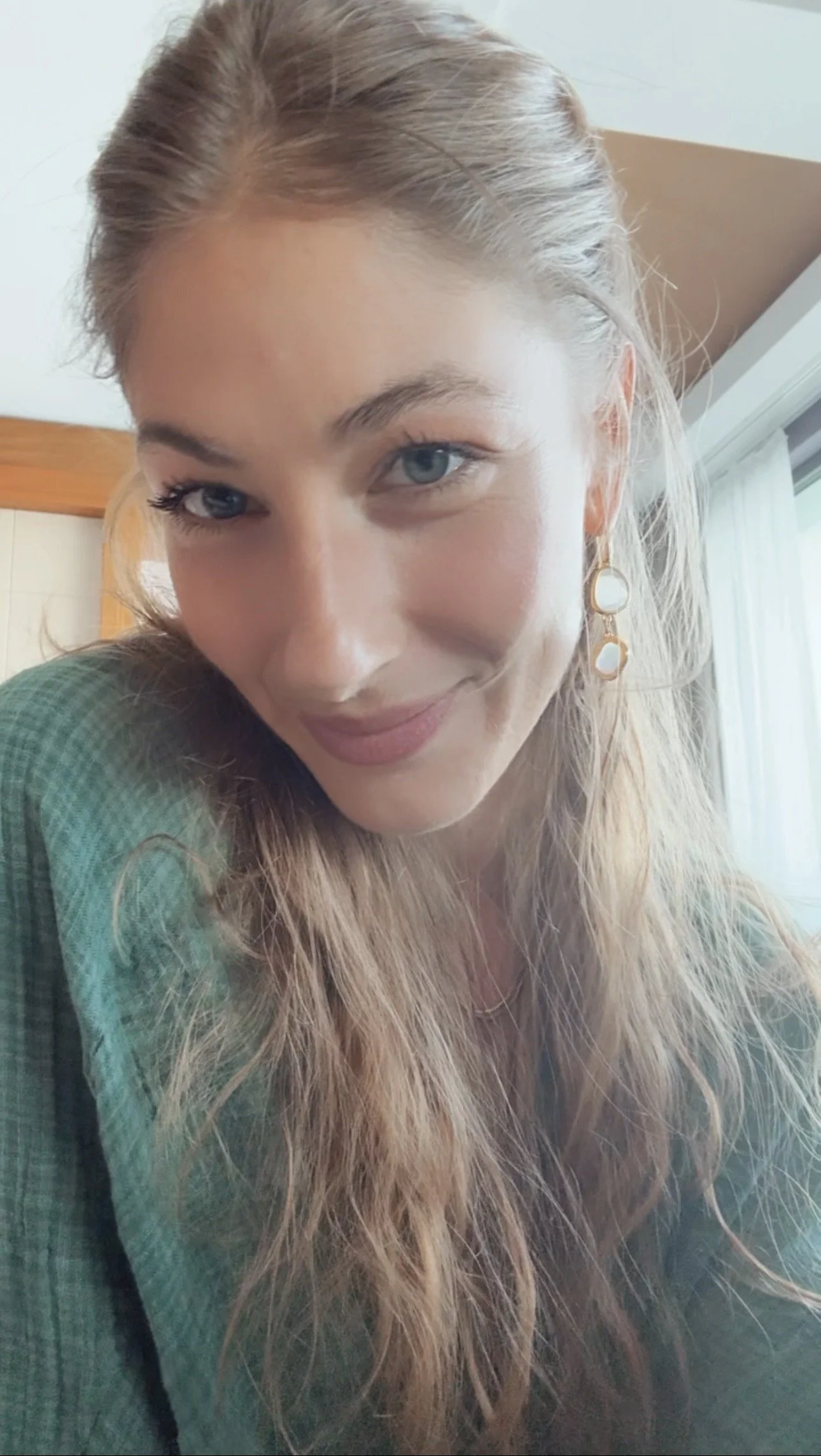 Close-up of a smiling woman with long, wavy blonde hair, blue eyes, and gold earrings, wearing a green top, indoors near a window.
