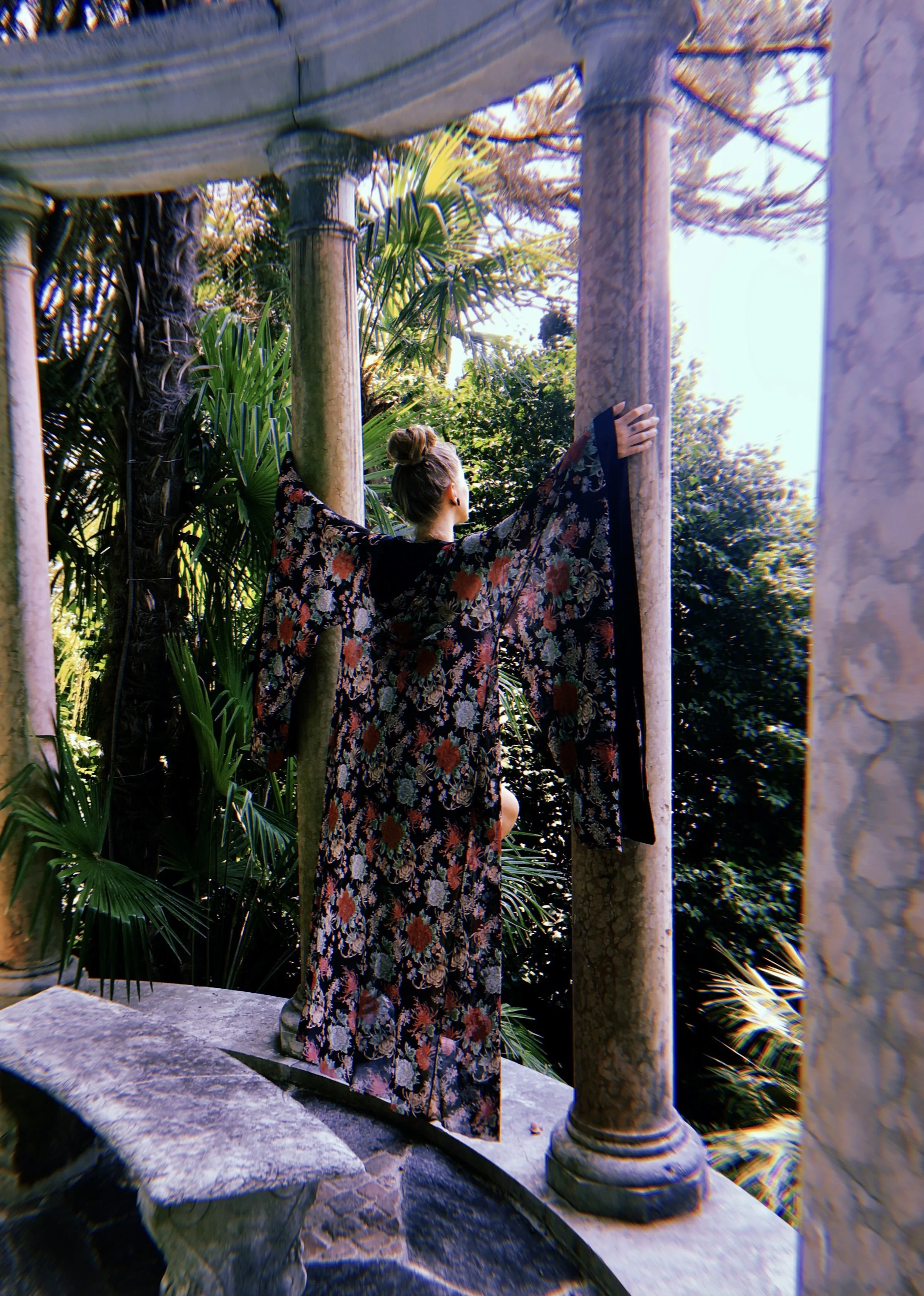 Woman in a floral kimono standing on stone platform with arms outstretched between Greek-style columns, lush greenery and palm trees in background.