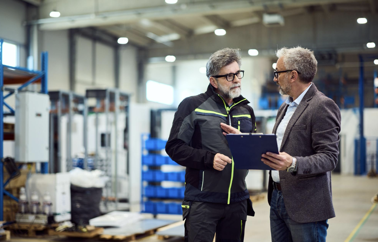 Two middle-aged men having a conversation in a warehouse, one holding a clipboard, surrounded by industrial shelves and equipment.