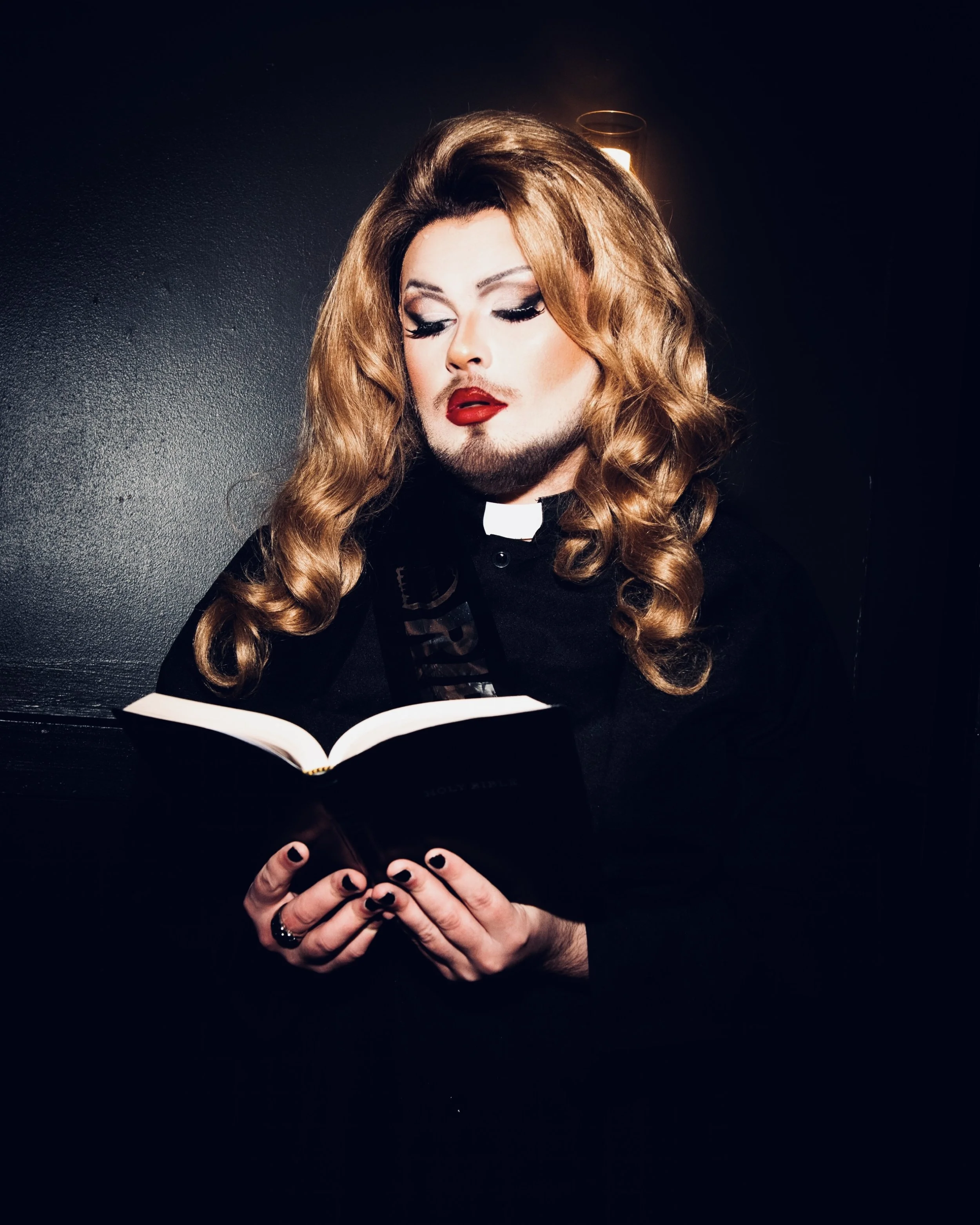 Blonde drag queen dressed as a reverend and reading from a bible.