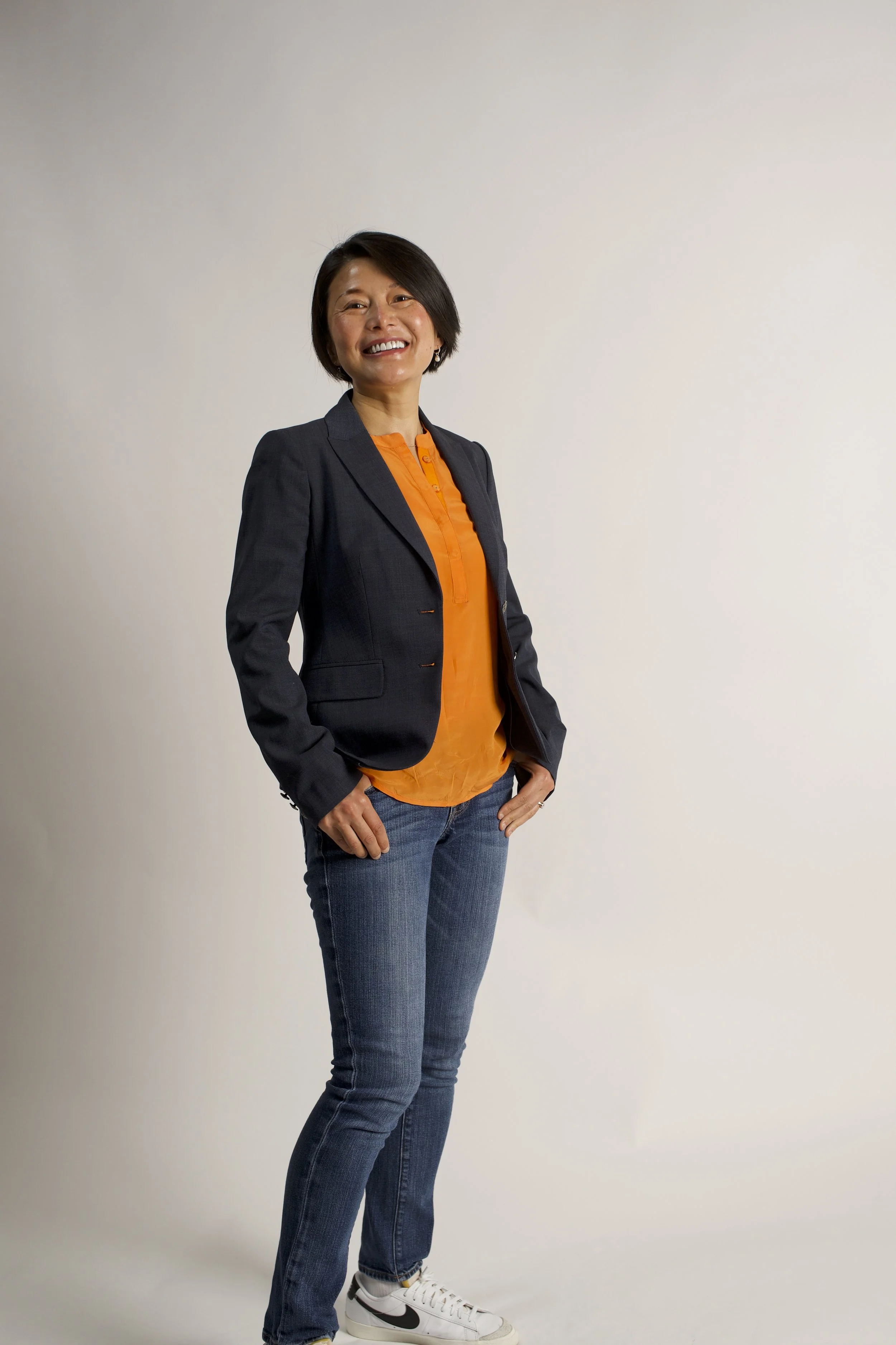A woman smiling, wearing a dark blazer over an orange shirt, with jeans and white sneakers, standing with her hands near her hips against a plain light background.